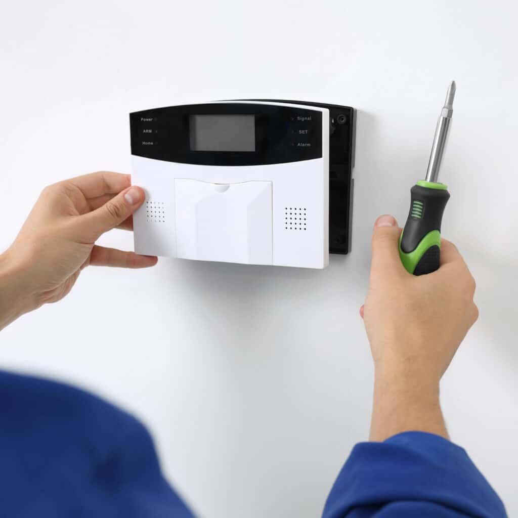 Installation alarme sécurité : technicien pose panneau contrôle Un technicien installe un panneau de contrôle d'alarme de sécurité blanc sur un mur blanc, tenant un tournevis. Textes: Power, Signal, ARM, SET, Home, Alarm.