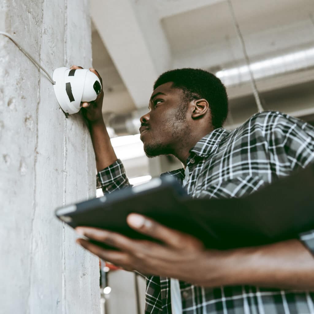Installation Caméra Sécurité | Solutions Professionnelles Jeune homme installant une caméra de sécurité sur un mur en béton texturé, consultant une tablette numérique.