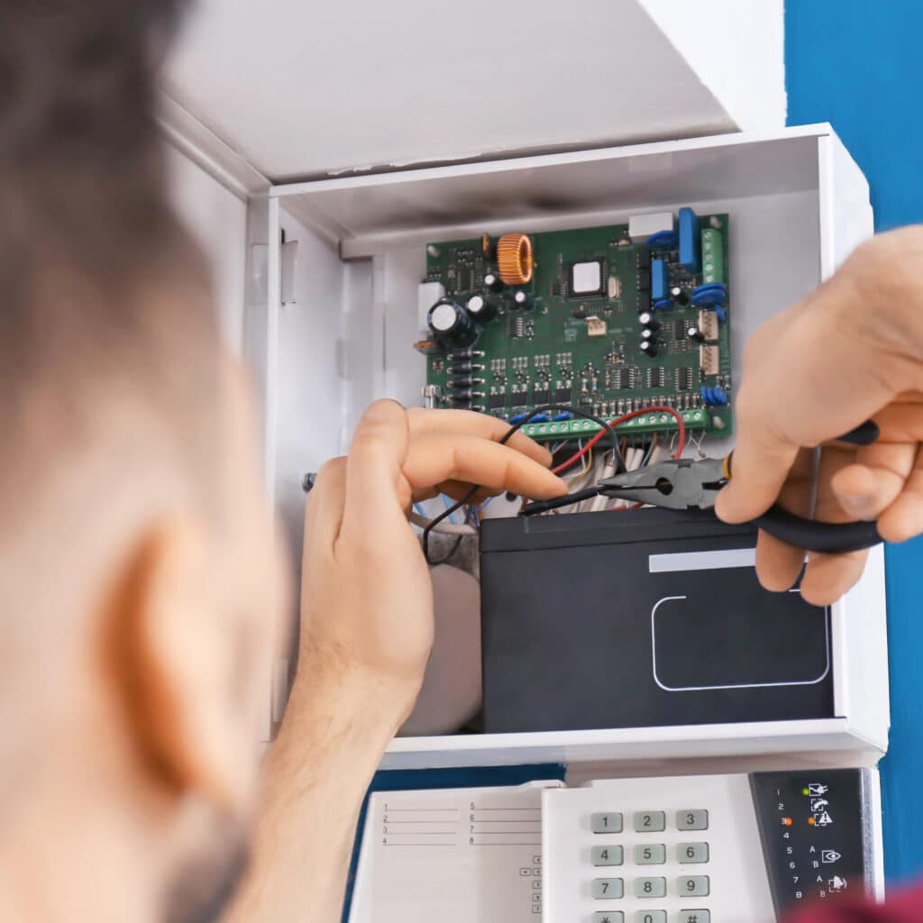 Technicien répare système alarme. Mains avec pince, fils, circuit imprimé, batterie. Clavier avec voyant d'erreur orange.