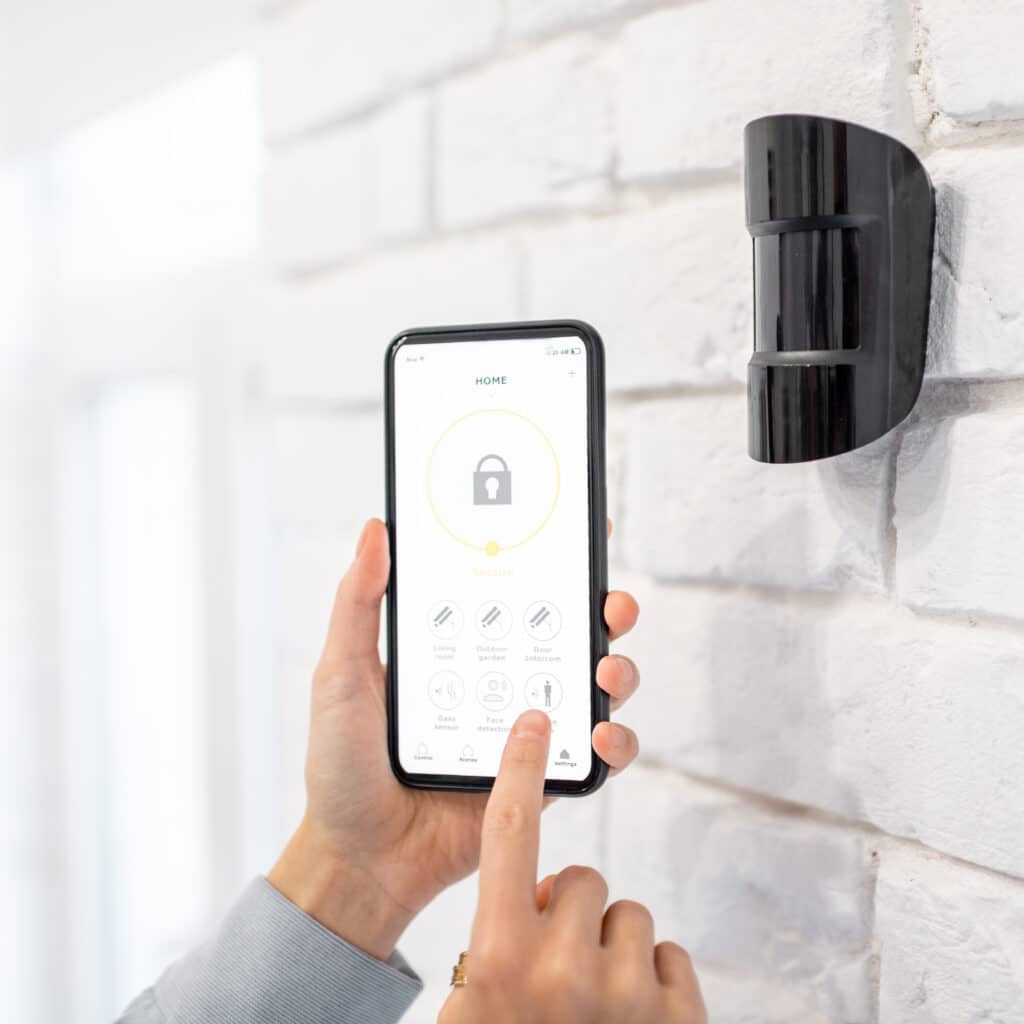 Main tapant appli "HOME Secure" sur smartphone pour contrôler divers capteurs (mouvement, gaz, visage). Capteur noir sur mur briques blanches.