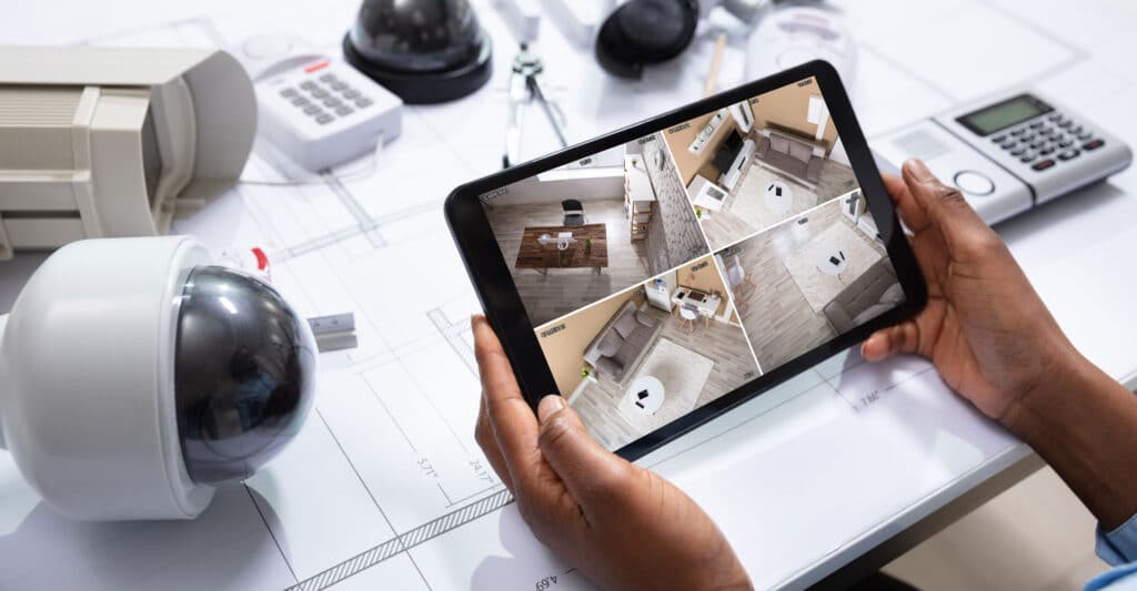 Main surveillant avec tablette, montrant 4 flux de caméras de sécurité intérieures sur des plans. Caméras et clavier d'alarme.